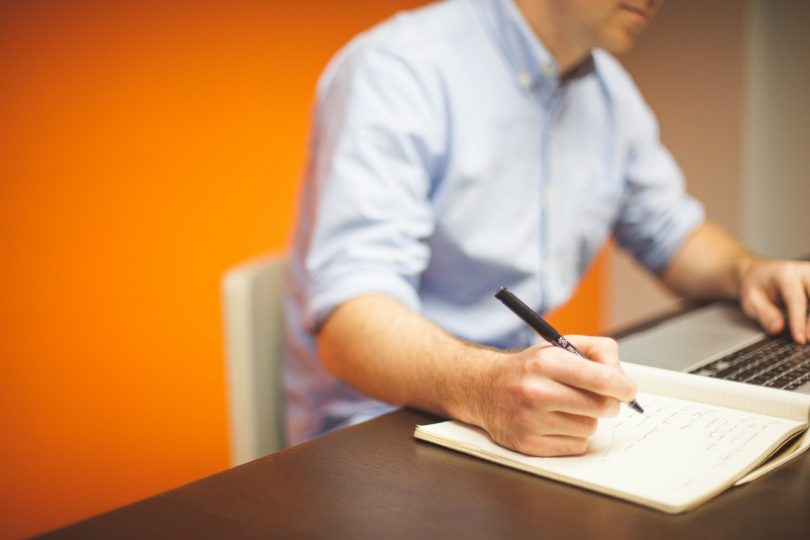 Person writing notes while looking at a laptop