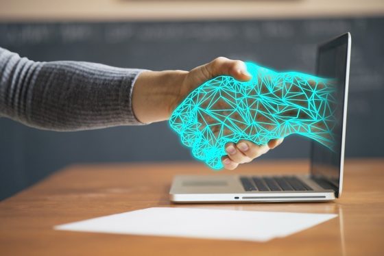 A wireframe hand extends from a laptop screen and shakes a human hand.
