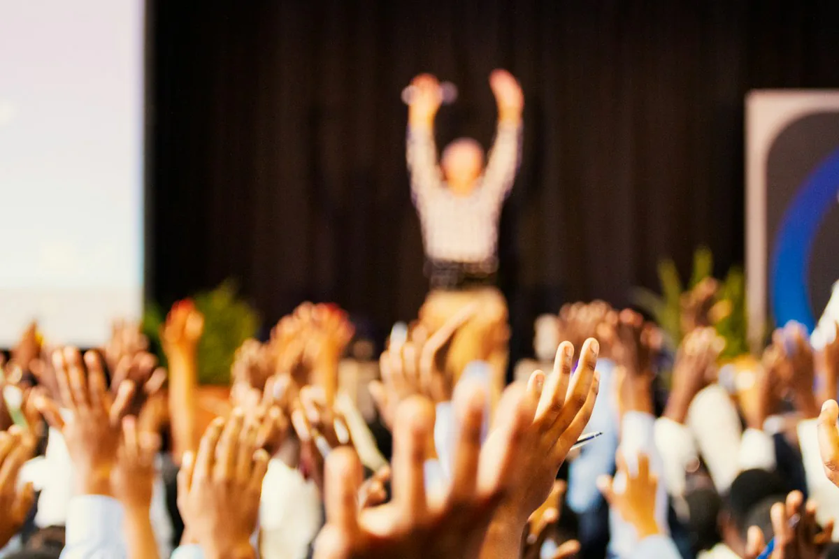 Raised hands in warm light during a conference, with a presenter softly focused in the background.