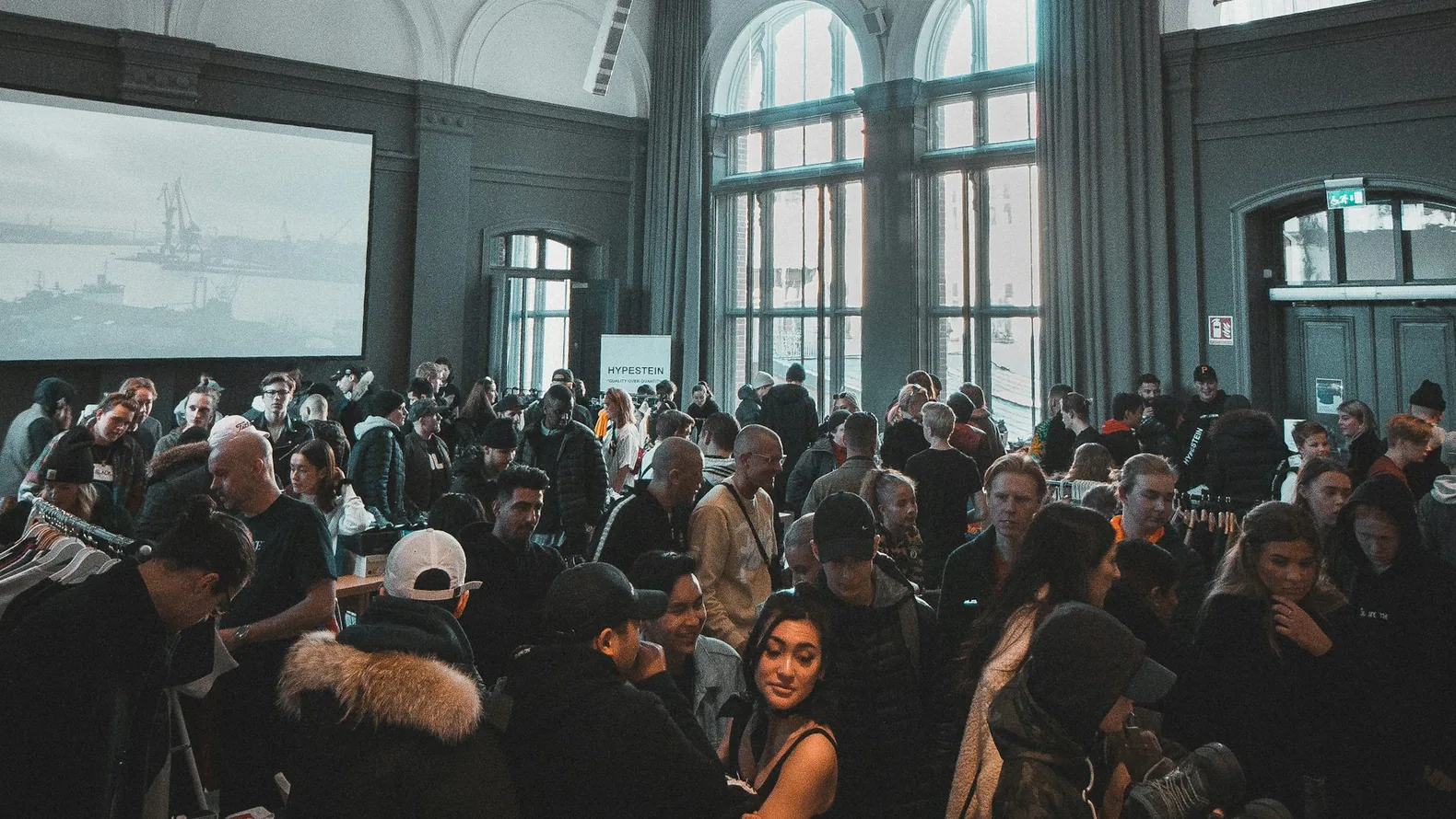 People gathering in a spacious hall lit by tall windows.