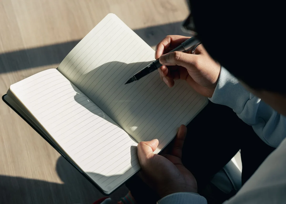 A person writes in an open notebook in warm natural window light.
