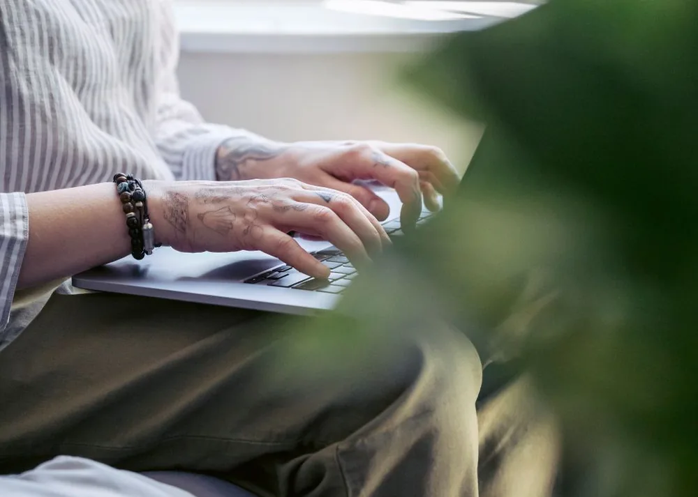 Hands working on a laptop beside a softly lit window.